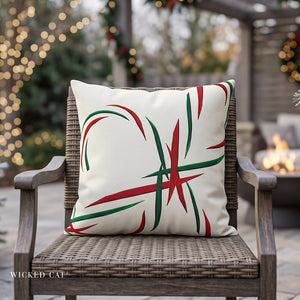 A square off-white outdoor throw pillow with a red and green abstract candy cane design, resting on a brown woven patio chair in front of a blurred outdoor fire pit and holiday lights background. The brand name 'WICKED CAT' is visible in the bottom left corner.