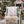A white wooden chair in a festive outdoor holiday setting, styled with a cream faux fur throw, a green knit blanket, and a white pillow printed with the word 'HOHOHO' in red and white candy cane patterns. A Christmas tree and a small 'Santa's Workshop' sign are blurred in the background.