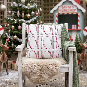 A white wooden chair in a festive outdoor holiday setting, styled with a cream faux fur throw, a green knit blanket, and a white pillow printed with the word 'HOHOHO' in red and white candy cane patterns. A Christmas tree and a small 'Santa's Workshop' sign are blurred in the background.