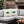 Two cream-colored outdoor throw pillows with green holly leaves and small pink berries, displayed on a gray sectional sofa in a cozy patio setting with a Christmas tree and fireplace in the background. The brand name 'WICKED CAT' is visible in the bottom left corner.