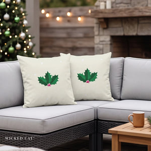 Two cream-colored outdoor throw pillows with green holly leaves and small pink berries, displayed on a gray sectional sofa in a cozy patio setting with a Christmas tree and fireplace in the background. The brand name 'WICKED CAT' is visible in the bottom left corner.