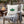A square off-white throw pillow with a green holly leaf and small pink berries printed on the front, resting on a wooden outdoor patio chair next to a gray plaid throw blanket. The scene is set in a cozy backyard with a fire pit, warm lights, and a Christmas tree in the background.
