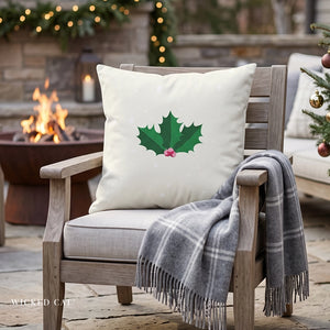 A square off-white throw pillow with a green holly leaf and small pink berries printed on the front, resting on a wooden outdoor patio chair next to a gray plaid throw blanket. The scene is set in a cozy backyard with a fire pit, warm lights, and a Christmas tree in the background.