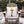 A square white throw pillow featuring a graphic of a single leg wearing a yellow lampshade and black high heels, resting on a snow-dusted wooden Adirondack chair in a snowy outdoor setting with Christmas trees in the background.