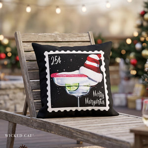 A square black throw pillow with a postage stamp design featuring a Christmas-themed margarita, resting on a weathered wooden chair in a festive outdoor setting with blurred Christmas lights and a tree in the background.