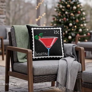 A square throw pillow with a vintage-style 25-cent stamp design featuring a red martini cocktail and the text 'MERRY MARTINI' and '25¢', styled on a gray wicker armchair with a green knit blanket and striped throw in a cozy, blurred holiday living room setting.