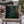 A square black throw pillow with a green holly and red berries Christmas tree design, resting on a wooden outdoor chair with a blurred festive patio background.