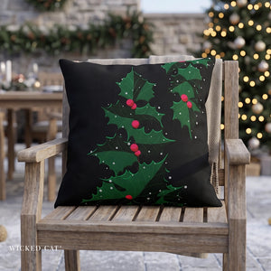 A square black throw pillow with a green holly and red berries Christmas tree design, resting on a wooden outdoor chair with a blurred festive patio background.