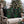 Two square black throw pillows with a green holly and red berries pattern, displayed on a beige outdoor patio chair in front of a decorated Christmas tree.