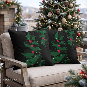 Two square black throw pillows with a green holly and red berries pattern, displayed on a beige outdoor patio chair in front of a decorated Christmas tree.
