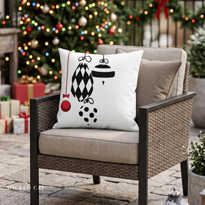 A white square throw pillow with a black holiday ornament graphic and a small red ornament accent, resting on a beige cushion in a wicker outdoor chair. The background shows a decorated Christmas tree with colorful lights and wrapped gifts.