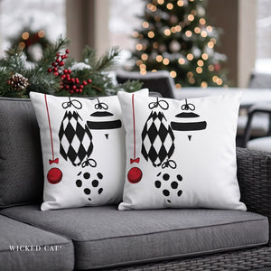 Two white square throw pillows with a black and red Christmas ornament pattern, displayed on a gray outdoor sofa with a blurred Christmas tree in the background.