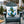 A square white outdoor throw pillow featuring a blue anchor design decorated with green holly leaves and red berries, displayed on a distressed teal Adirondack chair in a holiday-themed outdoor setting.