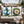 Two decorative outdoor throw pillows displayed on a wicker patio sofa. One pillow features a blue anchor with green holly leaves and red berries on a white background.