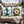 Two decorative outdoor throw pillows on a wicker patio sofa. One pillow features a blue nautical anchor wrapped in green holly with red berries on a white background.