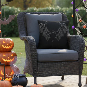 Gray wicker chair with a black bat pillow in an outdoor Halloween setting with pumpkins and decorations.