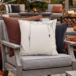 A white square throw pillow with a black spider and web design, styled on a gray outdoor patio chair with brown and navy accent pillows. The Wicked Cat brand logo is visible in the bottom left corner.