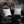 Two white square throw pillows with black spiderweb and spider designs, styled on a black leather sofa with a black faux fur throw blanket in a dark, decorated living room.