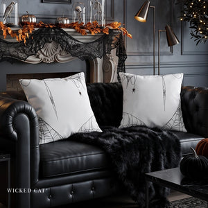 Two white square throw pillows with black spiderweb and spider designs, styled on a black leather sofa with a black faux fur throw blanket in a dark, decorated living room.