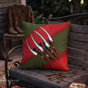 A square decorative throw pillow with a red and green background and a large white-and-brown claw graphic, placed on a wooden bench in an outdoor patio setting. A sweater, boots, and keys are visible around it, and the text 'WICKED CAT' appears in the lower left corner.
