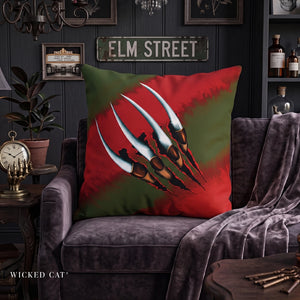 A square throw pillow featuring a red and green background with three metallic silver claw marks, styled on a dark purple velvet armchair in a themed room with an 'ELM STREET' sign and 'WICKED CAT' branding.