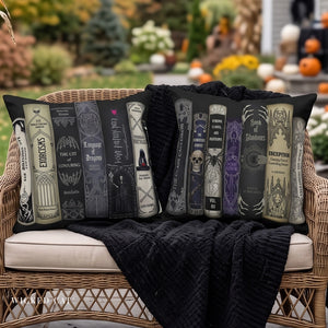 Two decorative throw pillows featuring gothic book spines, styled on a wicker chair with a black knit blanket in an outdoor garden setting.