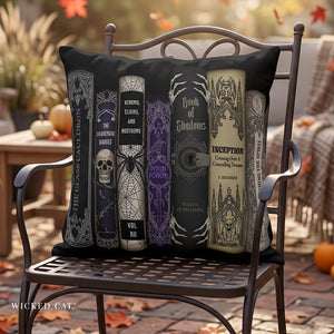 A decorative throw pillow shaped like a stack of gothic book spines sits on a wrought iron chair in an outdoor patio setting. The pillow features designs inspired by horror movies and pop culture, with black, gray, purple, and beige tones.