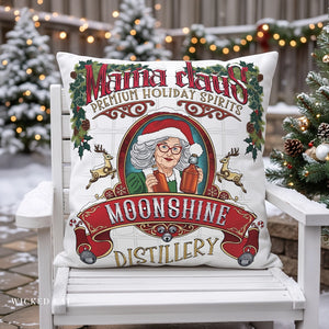 A square white decorative throw pillow with a colorful Christmas-themed print reading 'Mama claus Premium Holiday Spirits Moonshine Distillery,' displayed on a white outdoor chair with snowy trees and string lights in the background.