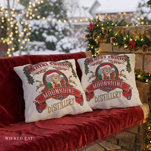 Two white decorative throw pillows with a festive Christmas 'Moonshine Distillery' design, displayed on a red velvet sofa in a decorated outdoor holiday setting.
