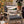 A wooden outdoor armchair with dark gray cushions is styled on a deck with Halloween decor, including a decorative throw pillow featuring a mummy eye design, pumpkins, a side table with a bottle, and a potted plant.