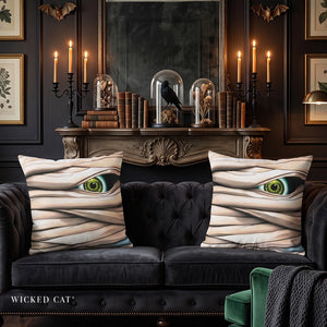 Two square throw pillows with a mummy eye print, styled on a black velvet sofa in a dark, gothic room with candles, books, and a mirror.