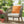 Chair with an orange diamond-patterned pillow on a patio with potted plants and flowers.