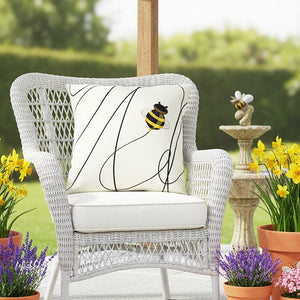 White wicker chair with a decorative pillow featuring a bee design, surrounded by potted plants and a garden setting.