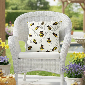 White wicker chair with a decorative pillow featuring bee patterns on a patio with plants and flowers.