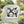 White wicker chair with a decorative pillow featuring flowers and a bee, set outdoors with greenery and flowers.