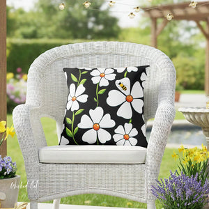 White wicker chair with a decorative pillow featuring flowers and a bee, set outdoors with greenery and flowers.