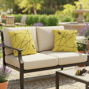 Outdoor patio set with beige cushions and yellow pillows featuring bee designs, surrounded by plants and a fountain.
