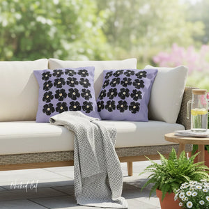Outdoor setting with a white sofa adorned with purple pillows featuring black floral patterns, a gray blanket, and a small table with a drink.