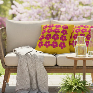 Outdoor setting with a beige sofa featuring yellow pillows with pink floral patterns, a wooden side table with glasses, and blurred floral background.