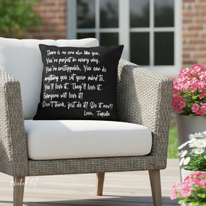 Black pillow with white text on a wicker chair outdoors with flowers in the background