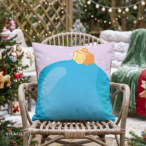 A square decorative throw pillow with a Christmas ornament design, displayed on a wicker chair in a festive outdoor holiday setting with a decorated tree and warm string lights in the background.
