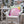 A pink and mint green decorative throw pillow with a big pink ornament  graphic, resting on a white wicker outdoor chair in a snowy patio.