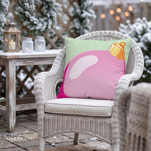 A pink and mint green decorative throw pillow with a big pink ornament  graphic, resting on a white wicker outdoor chair in a snowy patio.