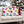 Two square throw pillows on a cream sofa in a holiday living room setting. The front pillows have a cream background with a colorful pattern of Christmas ornaments in pink, blue, purple, green, and yellow, with a small gold outline.