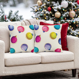 Two square throw pillows on a cream sofa in a holiday living room setting. The front pillows have a cream background with a colorful pattern of Christmas ornaments in pink, blue, purple, green, and yellow, with a small gold outline.
