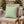 A square decorative throw pillow in sage green with a pink reindeer face and white snowflake pattern, resting on a woven outdoor chair in a cozy holiday setting with Christmas trees and warm lights in the background.
