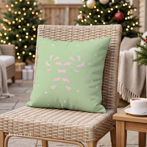 A square decorative throw pillow in sage green with a pink reindeer face and white snowflake pattern, resting on a woven outdoor chair in a cozy holiday setting with Christmas trees and warm lights in the background.