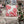 A square outdoor throw pillow with a red and white candy cane swirl pattern on a white background with tiny black polka dots, displayed on a distressed white metal patio chair in a festive holiday setting.