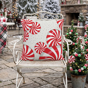 A square outdoor throw pillow with a red and white candy cane swirl pattern on a white background with tiny black polka dots, displayed on a distressed white metal patio chair in a festive holiday setting.