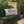 Two white square throw pillows with a pink and green candy cane and holly design, resting on a green fringed throw blanket on a wicker sofa in an outdoor patio setting.